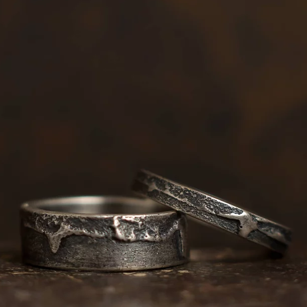 A close-up of two rustic, metal rings against a dark, blurred background. The handmade Wide Ribbon Ring by Septentrion boasts a rugged, textured appearance with unique patterns and slightly weathered, vintage look. One ring is lying flat while the other is propped up against it, showcasing their distinctive charm.