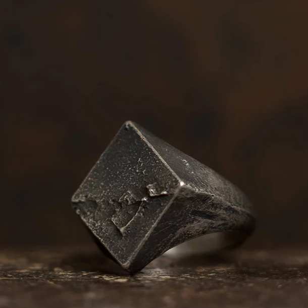A close-up of a dark metallic ring crafted from patinated 925 silver, with a wide square surface featuring rough, textured details. The Diamond Signet Ring by Septentrion, resembling an aged and weathered diamond signet ring, is set against a blurred brownish background.