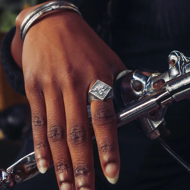 Close-up of a person's hand resting on the handlebar of a motorcycle. The hand is adorned with handmade jewelry, including a **Septentrion Diamond Signet Ring** and a patinated 925 silver bracelet. The motorcycle handlebar features shiny, reflective metal parts.