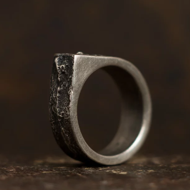 A close-up shot of a rugged, patinated 925 silver ring with an irregular, angular design, set against a dark background. This handmade jewellery piece by Septentrion features a rough, textured exterior that contrasts beautifully with its smooth, circular band.