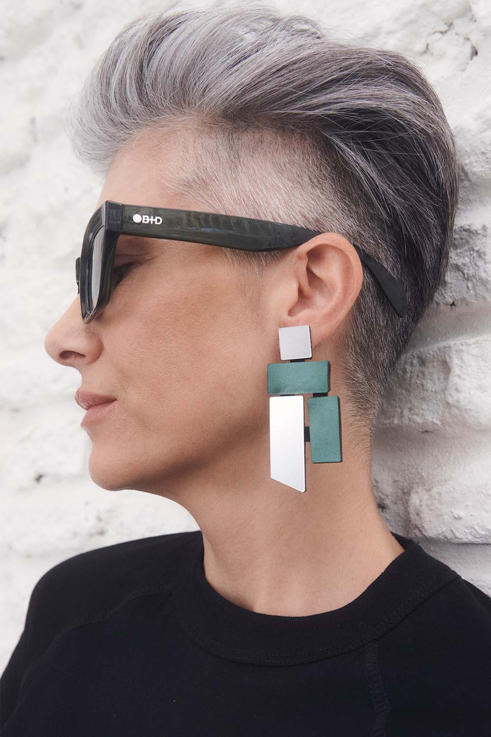 A person with short gray hair and sunglasses is shown in profile against a white wall, wearing Iskin Sisters' Piet Silver & Green Earrings—bold, geometric jewelry inspired by Mondrian with a modern touch.