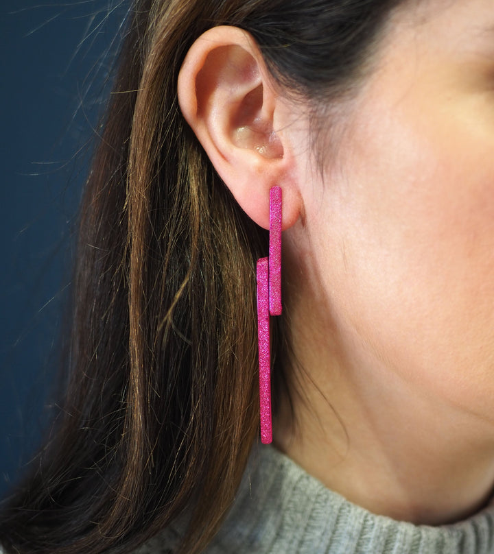 Close-up of a woman wearing Lysithea Magenta Pink Earrings by Theodora D. The handcrafted earrings consist of two elongated rectangular pieces that hang vertically from her earlobe. The woman has long brown hair and is wearing a light grey top. The background is dark.