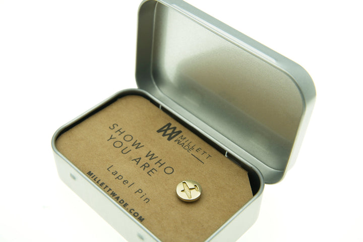 A small brass Plane Lapel Pin displayed in an open rectangular metal tin. The pin features a minimalist design and is attached to a piece of cardboard that reads "SHOW WHO YOU ARE" and "Plane Lapel Pin." The brand name "Millett Wade" and website "millettwade.com" are also visible.