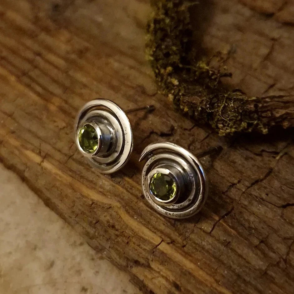 A pair of sterling silver stud earrings with green gemstones in the center, displayed on a rustic wooden surface with a piece of moss. The Peridot Sun Seeker Spiral Stud Earrings by Caroline Stokesberry-lee have concentric circular designs, enhancing the elegant and earthy aesthetic reminiscent of ancient Ireland.