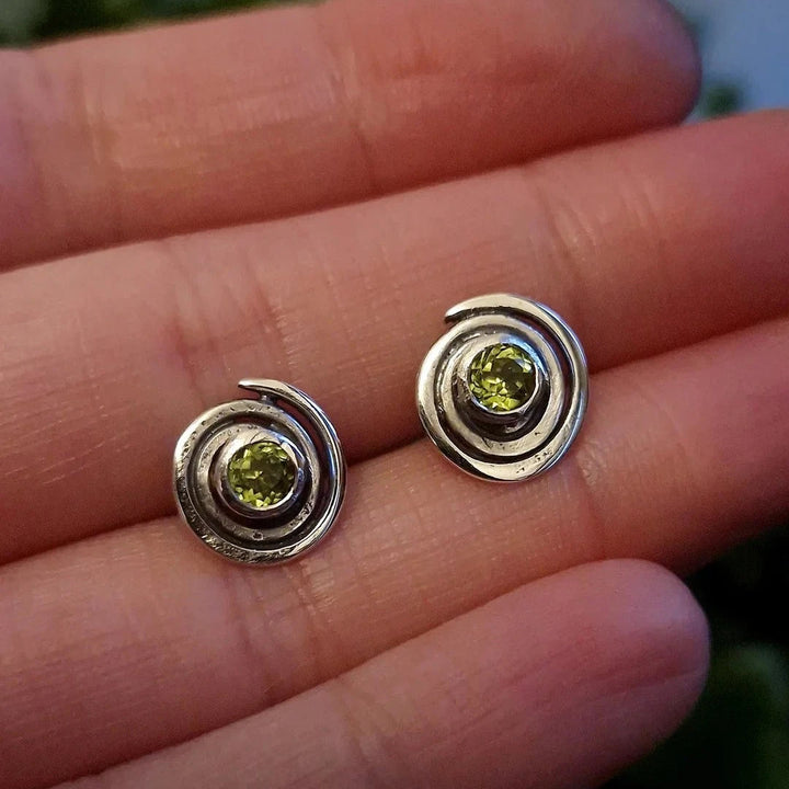 A hand holding a pair of Peridot Sun Seeker Spiral Stud Earrings from Caroline Stokesberry-lee made of sterling silver. Each earring showcases a spiral design reminiscent of ancient Ireland, with a green gemstone set in the center. The background is slightly blurred, emphasizing the detailed craftsmanship of the earrings.