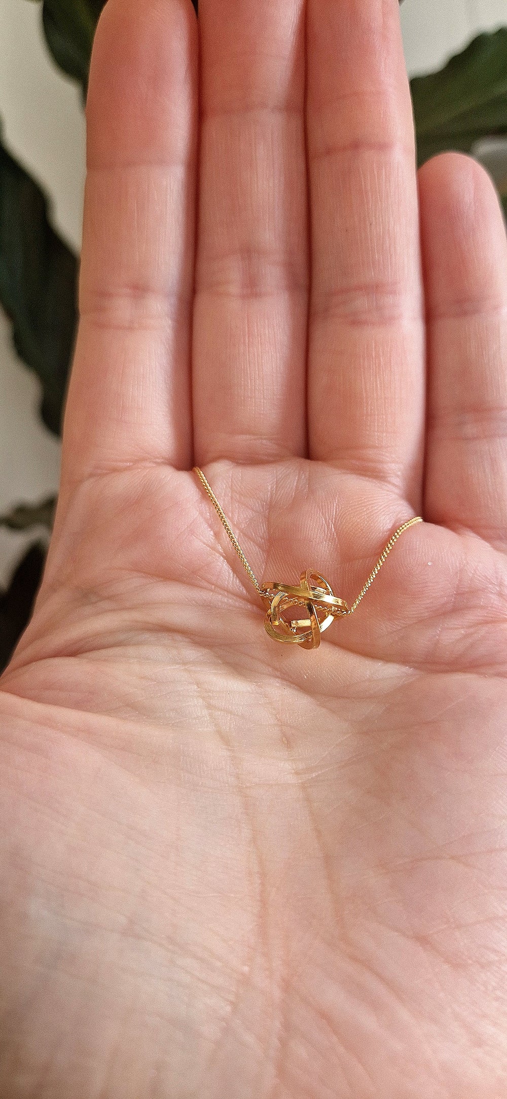 A close-up shows a hand holding the Caroline Stokesberry-lee Knot Pendant Gold, a handmade necklace delicately crafted in 18ct yellow gold plated metal. The open palm is set against a softly blurred backdrop with green leaves.