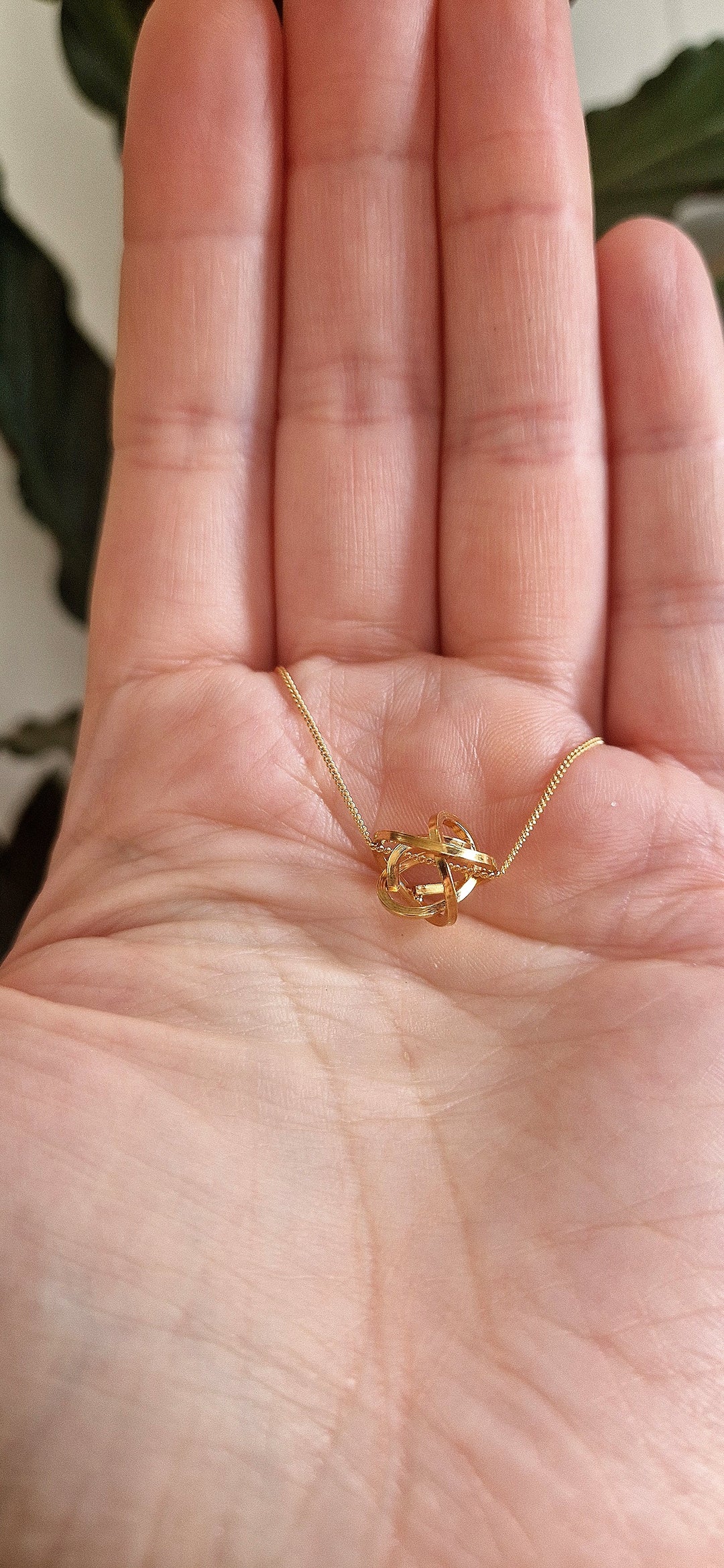A close-up shows a hand holding the Caroline Stokesberry-lee Knot Pendant Gold, a handmade necklace delicately crafted in 18ct yellow gold plated metal. The open palm is set against a softly blurred backdrop with green leaves.