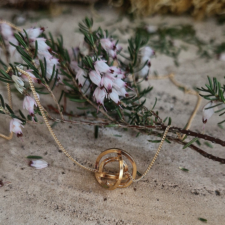 The Knot Pendant Gold by Caroline Stokesberry-lee, an 18ct yellow gold plated geometric necklace, is displayed on sand next to pink and white bell-shaped flowers, highlighting its natural elegance.