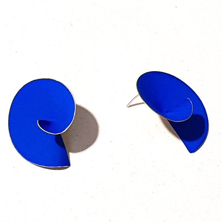 A pair of modern, handmade Matt Swirl Earrings by Georgia Charal in a vibrant cobalt blue pigment and gold-plated silver lies on a white background. Each earring has a unique sculptural design resembling a curled or twisted shape.