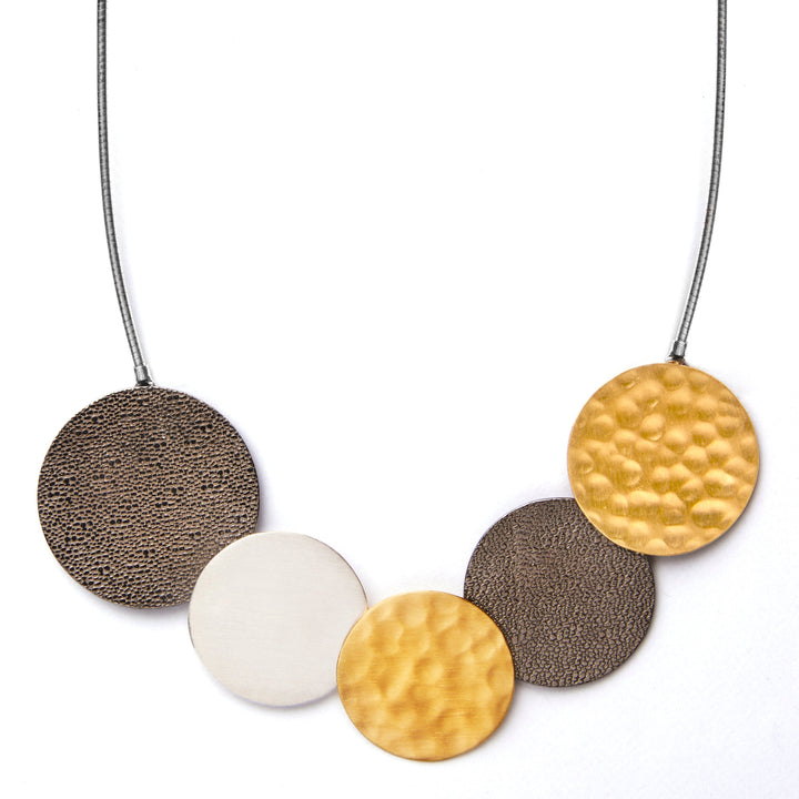 A Portobello Tricolour Necklace from The Collective Dublin featuring five circular pendants. The leftmost and second from the right pendants have a textured dark brown surface, the middle pendant is smooth and silver, and the first and last pendants are gold plated silver. The 16-inch choker attaches them to a thin cord.