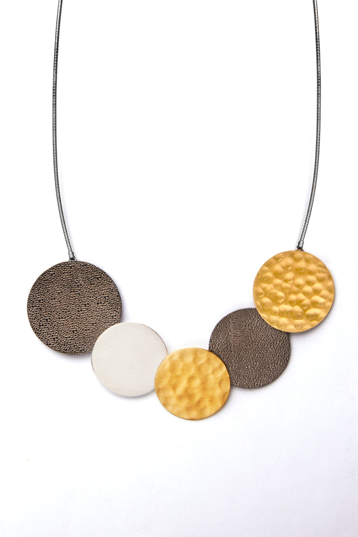 The Collective Dublin's Portobello Tricolour Necklace features a 16 inch sterling silver choker with a chain holding five large, round pendants. From left to right, the pendants are textured dark brown, hammered gold plated silver, smooth sterling silver, hammered gold plated silver, and textured dark brown, creating a symmetric pattern.