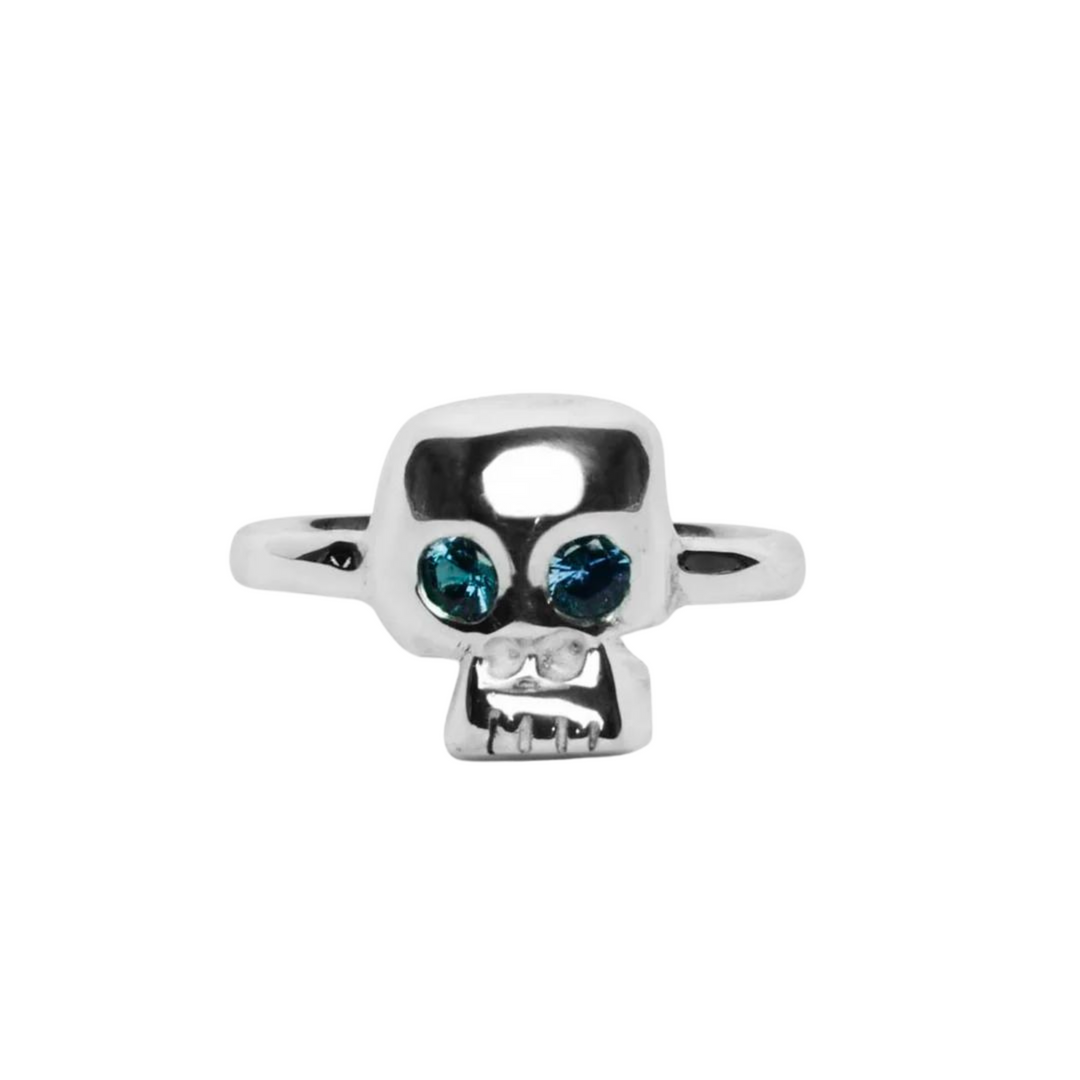 The Silver Memento Mori Skull Ring by Cosmic Boulevard features a sterling silver ring with a skull design that has two blue gemstones as eyes. The skull has a stylized appearance with smooth contours and a slightly exaggerated facial expression, embodying the essence of memento mori. The band of this handmade jewellery piece is simple and unadorned.