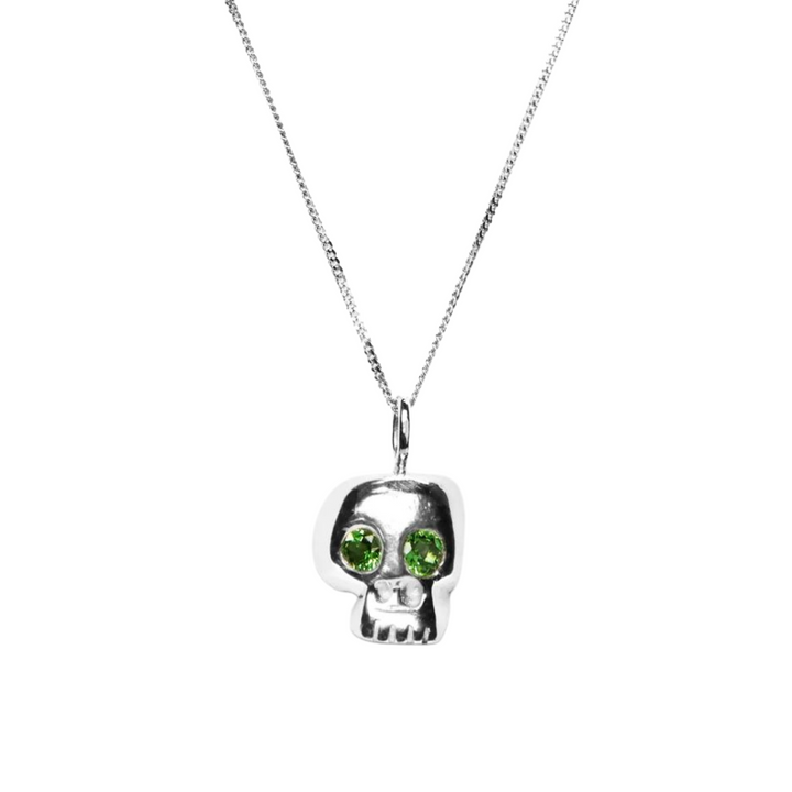 A Silver Memento Mori Skull Pendant from Cosmic Boulevard, featuring a pendant with two large, round green gemstones for eyes. The chain is thin and delicate, set against a plain white background. Handmade jewellery at its finest.