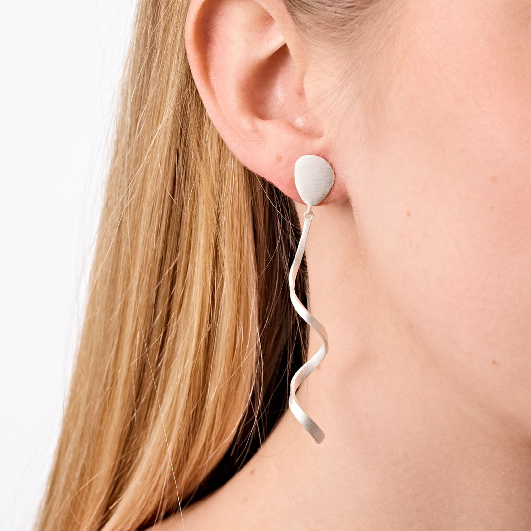Close-up of a woman’s ear adorned with Tezer’s Spiral Twist Earrings. These modern sterling silver earrings feature a circular stud and a long, wavy spiral-shaped drop. The woman’s straight, light blonde hair frames the elegant design.