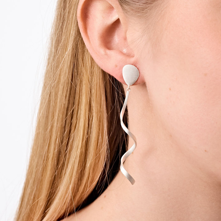 Close-up of a woman’s ear adorned with Tezer’s Spiral Twist Earrings. These modern sterling silver earrings feature a circular stud and a long, wavy spiral-shaped drop. The woman’s straight, light blonde hair frames the elegant design.