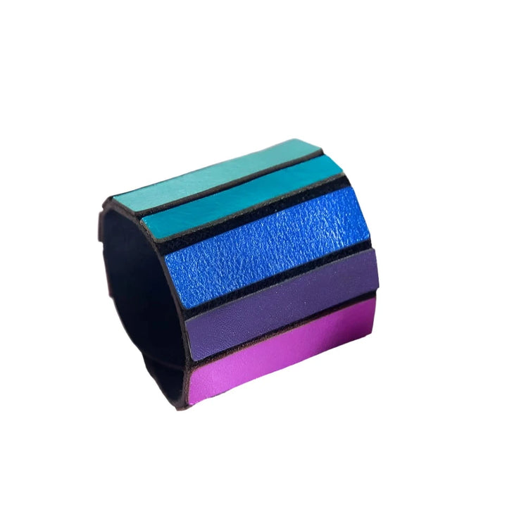 The Rainbow Round Bracelet by Iskin Sisters, a wide leather cuff adorned with horizontal stripes in vibrant shades of teal, blue, purple, and pink, is displayed against a geometric white background. The bracelet's striking colors stand out vividly against the clean, angular lines of the folded paper beneath it.