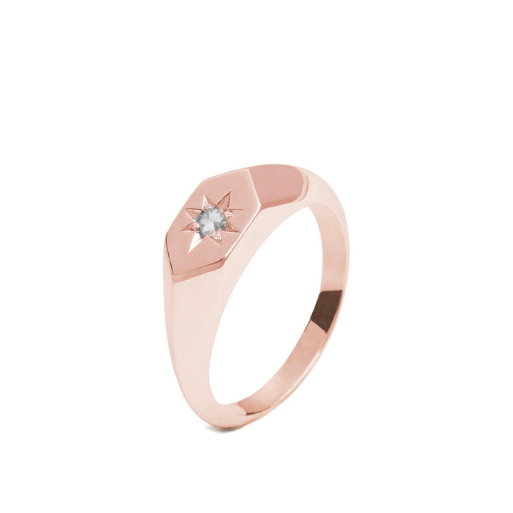 The Starlight Diamond Birthstone 9ct Gold Signet Ring by Cosmic Boulevard is a minimalist rose gold ring with a hexagonal-shaped top featuring a small, centrally-set diamond, the birthstone of April. Handmade in Dublin, the ring has a smooth, polished finish and a modern design, blending simplicity with elegance.