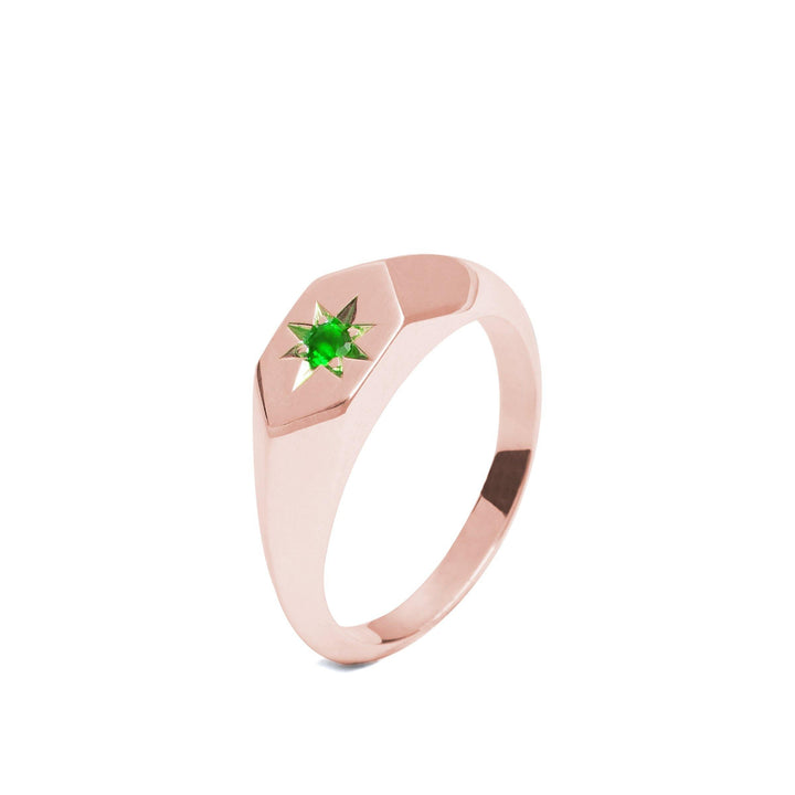 A rose gold Starlight Emerald Birthstone 9ct Gold Signet Ring by Cosmic Boulevard with a hexagonal face featuring a star-shaped setting holds the birthstone of May, a small green gemstone at its center. Handmade in Dublin, the ring is beautifully displayed against a plain white background.