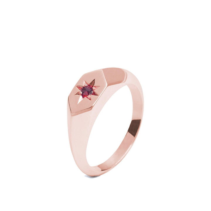 A sleek, modern rose gold ring features a star-shaped setting that holds a small red gemstone at its center. The band, with a smooth, polished finish and slightly angular shape near the setting, has the minimalistic yet elegant aesthetic of a Starlight Ruby Birthstone 9ct Yellow Gold Signet Ring from Cosmic Boulevard, making it perfect as the birthstone of July.