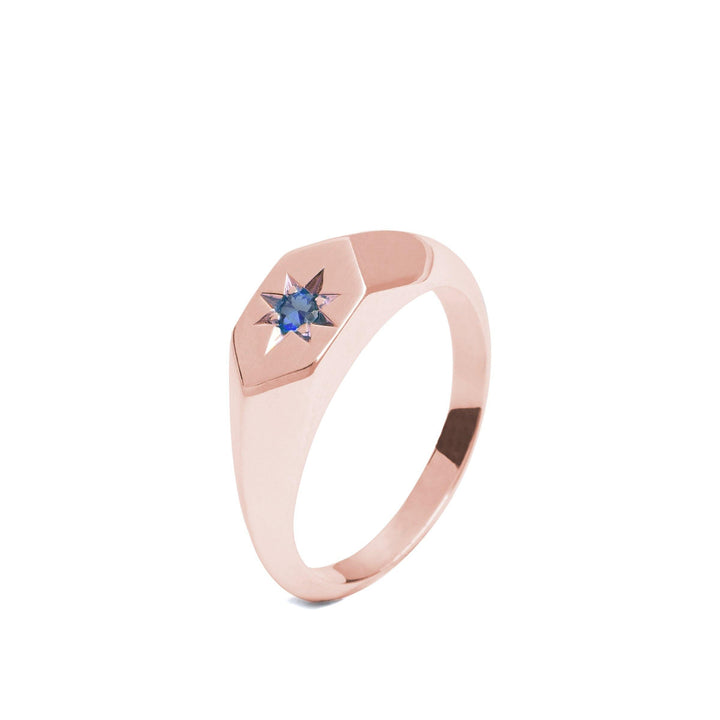 This rose gold ring, crafted from 9ct gold, boasts a hexagonal face with a star-shaped design set with a small blue sapphire gemstone in the center. The band is simple and polished, showcasing a minimalist and elegant Cosmic Boulevard Starlight Sapphire Birthstone 9ct Gold Signet Ring style perfect for September birthstones.