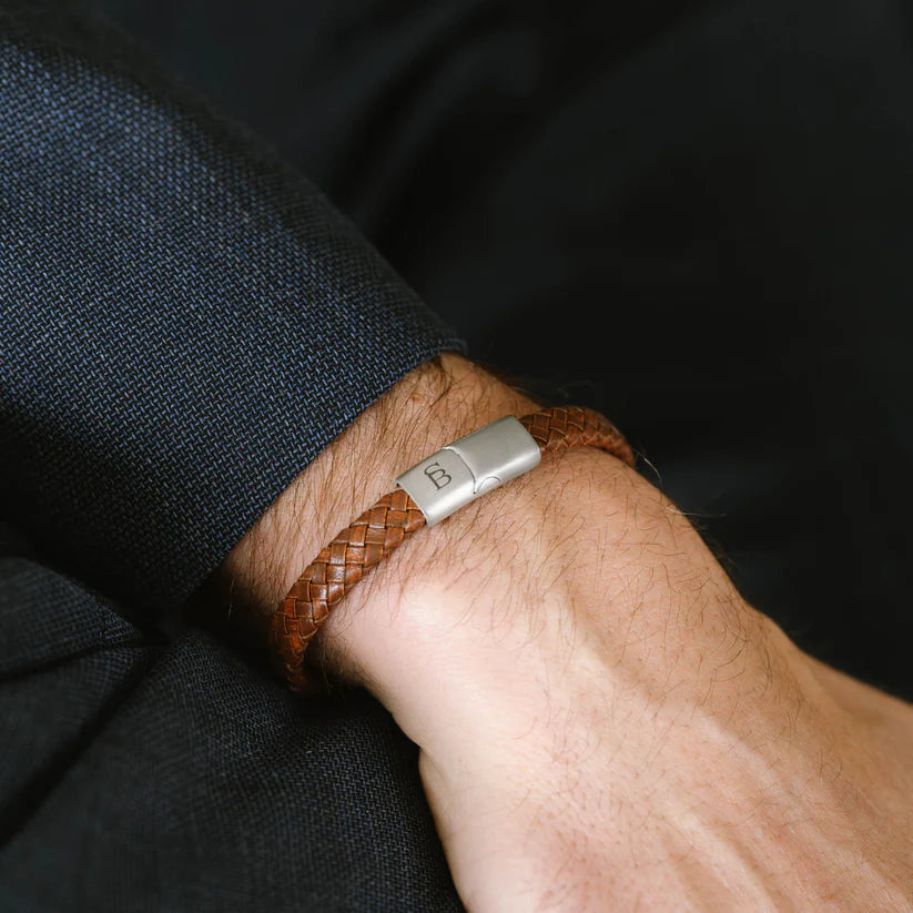 A close-up of a person's wrist adorned with the Riley - Caramel, a sturdy brown braided leather bracelet from Steel and Barnett, featuring a magnetic clasp engraved with the letter "B." The person is dressed in a dark-colored suit.