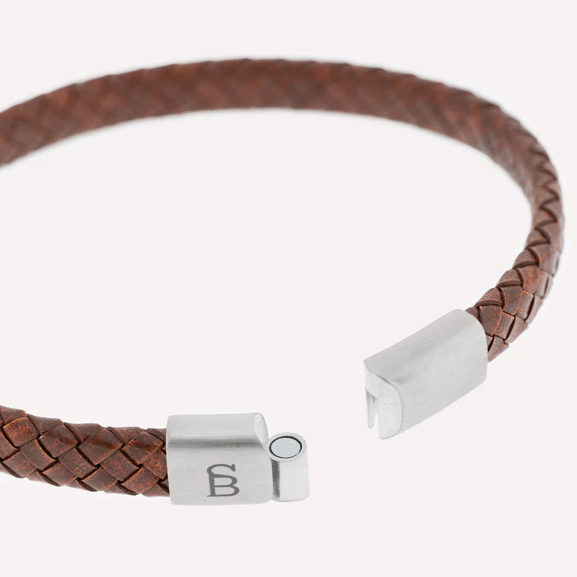 A close-up image of the Riley - Caramel braided leather bracelet by Steel and Barnett, featuring sturdy silver magnetic clasp ends. The clasp on the left has a metal cylinder, while the one on the right has a slot for the cylinder to fit in. One clasp is engraved with the Steel and Barnett logo.