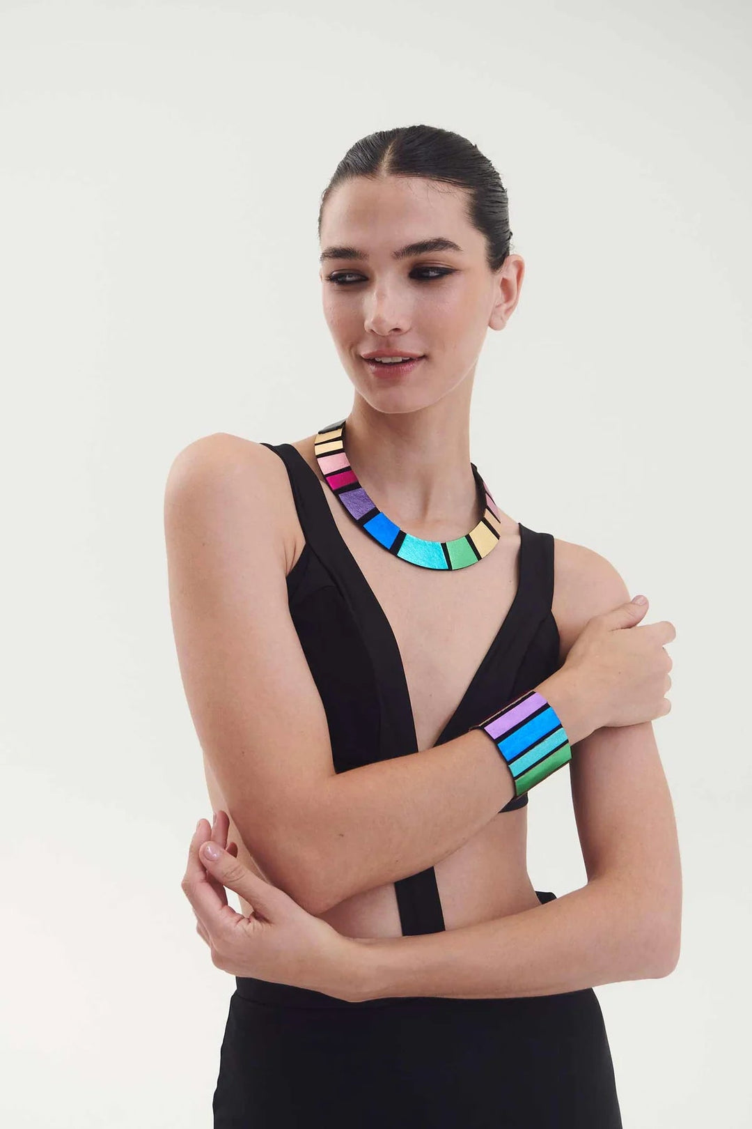 A person with slicked-back hair poses confidently, wearing a minimalistic black outfit that exposes their midriff. They accessorize with a colorful, geometric necklace and the Rainbow Round Bracelet by Iskin Sisters. The background is plain white.
