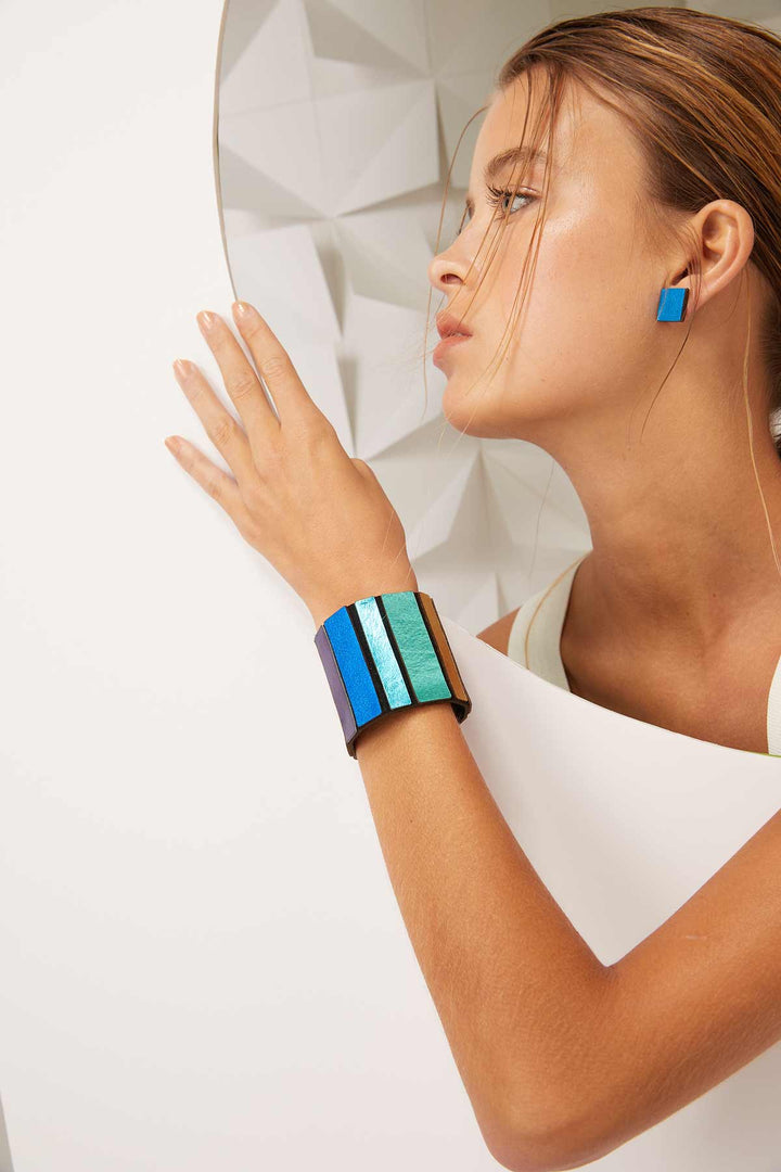 A woman with slicked-back hair models geometric blue and teal square earrings paired with the Rainbow Round Bracelet from Iskin Sisters. She is posed artistically against a white, textured geometric backdrop, highlighting the modern and colorful jewelry.