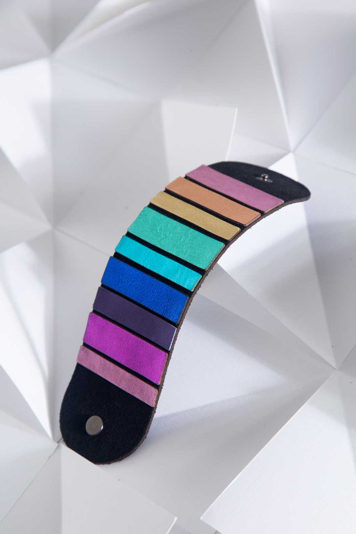 The Rainbow Round Bracelet from Iskin Sisters is featured against a geometric white background, showcasing its fan-shaped leather design. The bracelet displays strips in an array of colors including shades of purple, blue, teal, green, yellow, and pink. Each colorful strip is attached to a dark suede base with a metal button at one end.