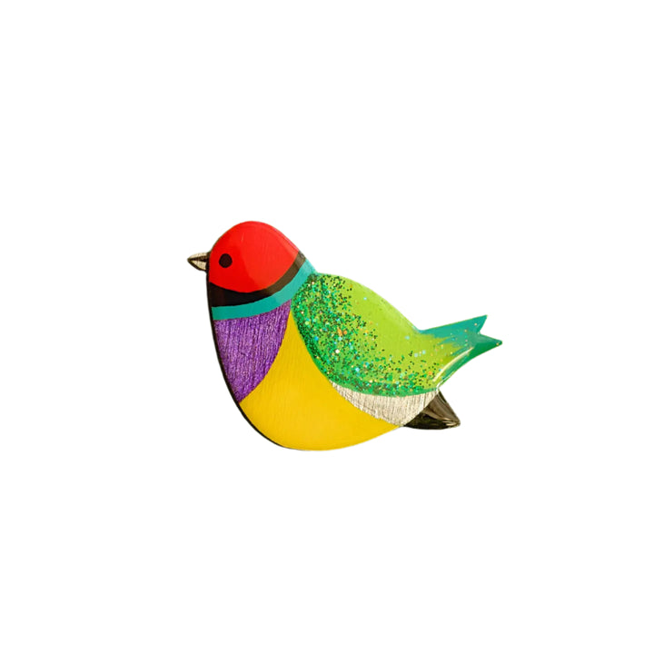The Naoi Rainbow Finch Pin Brooch features a white background and a vibrant design inspired by the Gouldian Finch, with a red head, green and yellow body, purple chest, green tail, and glitter accents on the back.