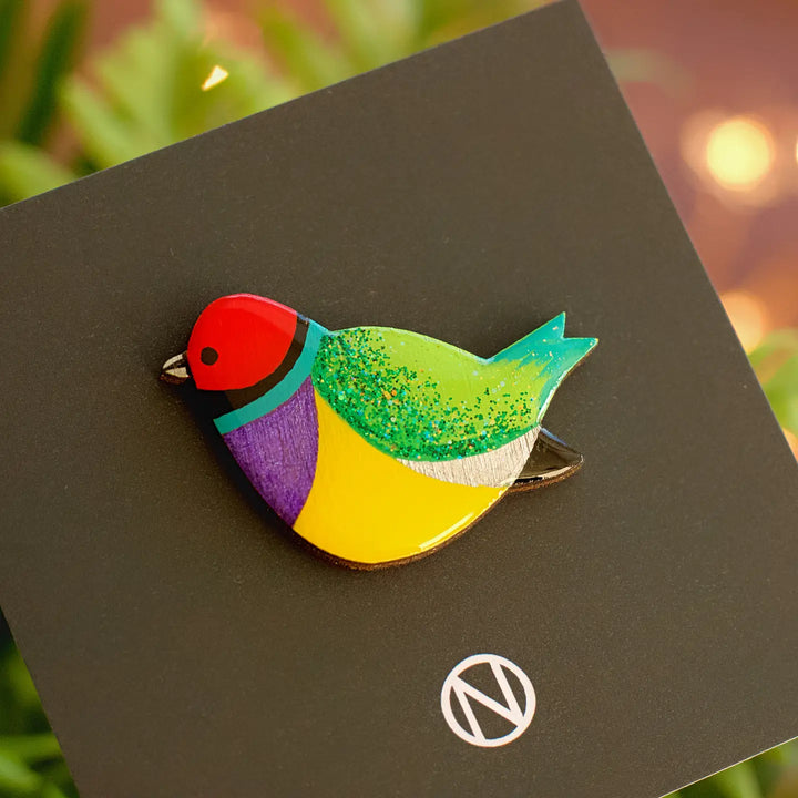 The Rainbow Finch Pin Brooch by Naoi is a vibrant, hand-painted wooden accessory with red, green, yellow, purple, and blue sections. Displayed on a black card with Naoi's "N" logo in a white circle; blurred green leaves are in the background.