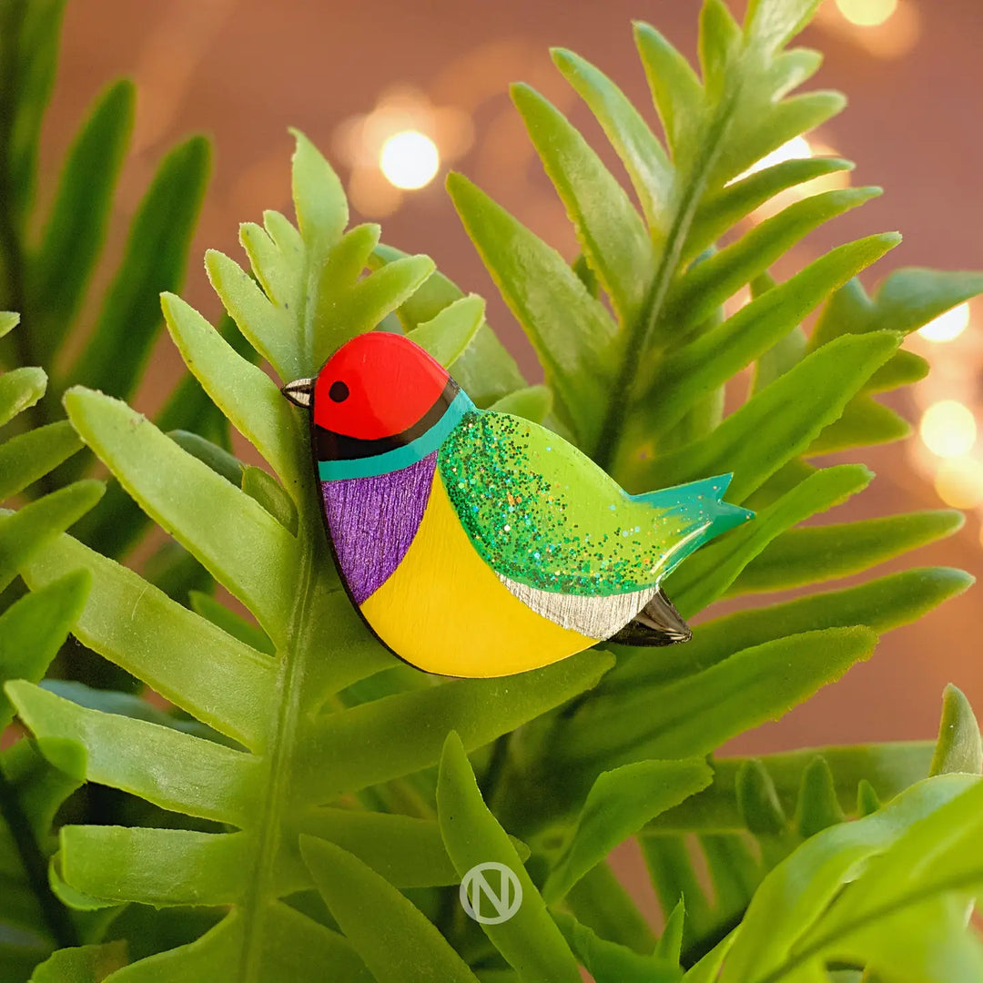 The Naoi Rainbow Finch Pin Brooch is a hand-painted wooden accessory inspired by the Gouldian Finch, featuring vibrant red, green, yellow, and purple hues as it rests on fern leaves with soft lights in the background.