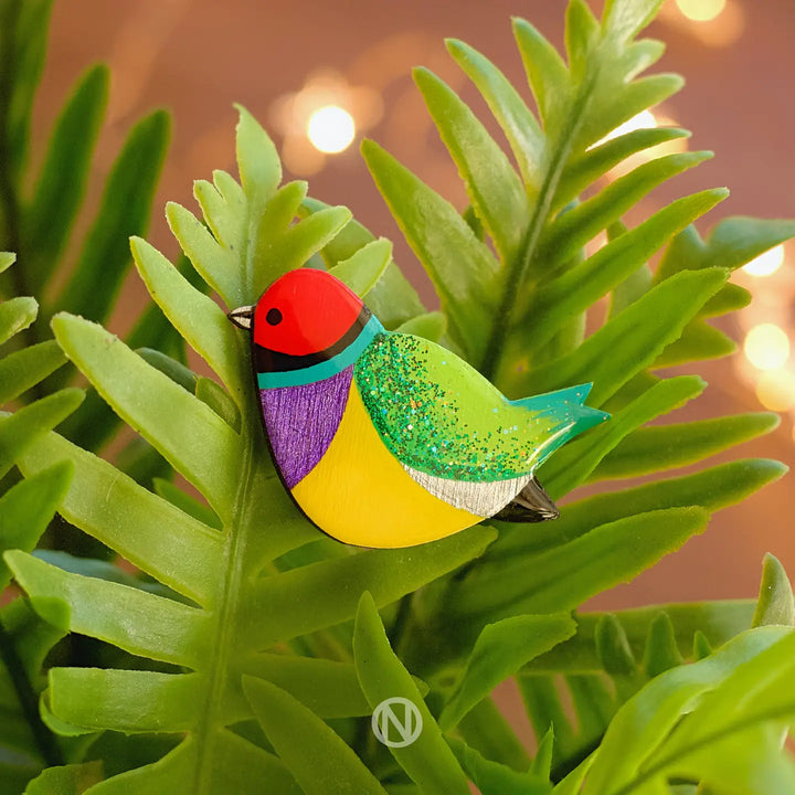 The Naoi Rainbow Finch Pin Brooch is a hand-painted wooden accessory inspired by the Gouldian Finch, featuring vibrant red, green, yellow, and purple hues as it rests on fern leaves with soft lights in the background.