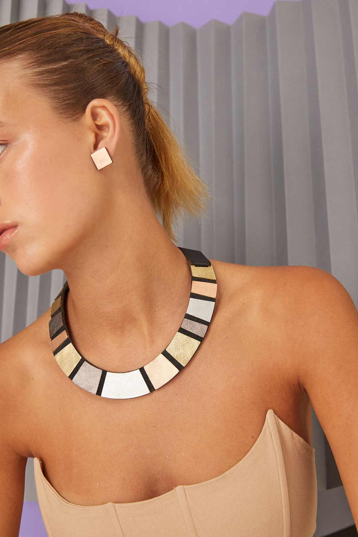 A person with slicked-back hair is wearing the Rainbow Leather Metallics Necklace by Iskin Sisters, which features chunky, metallic rectangular segments. They also have matching square stud earrings. The individual is dressed in a strapless beige top and is posing against a gray abstract backdrop with pleated details.