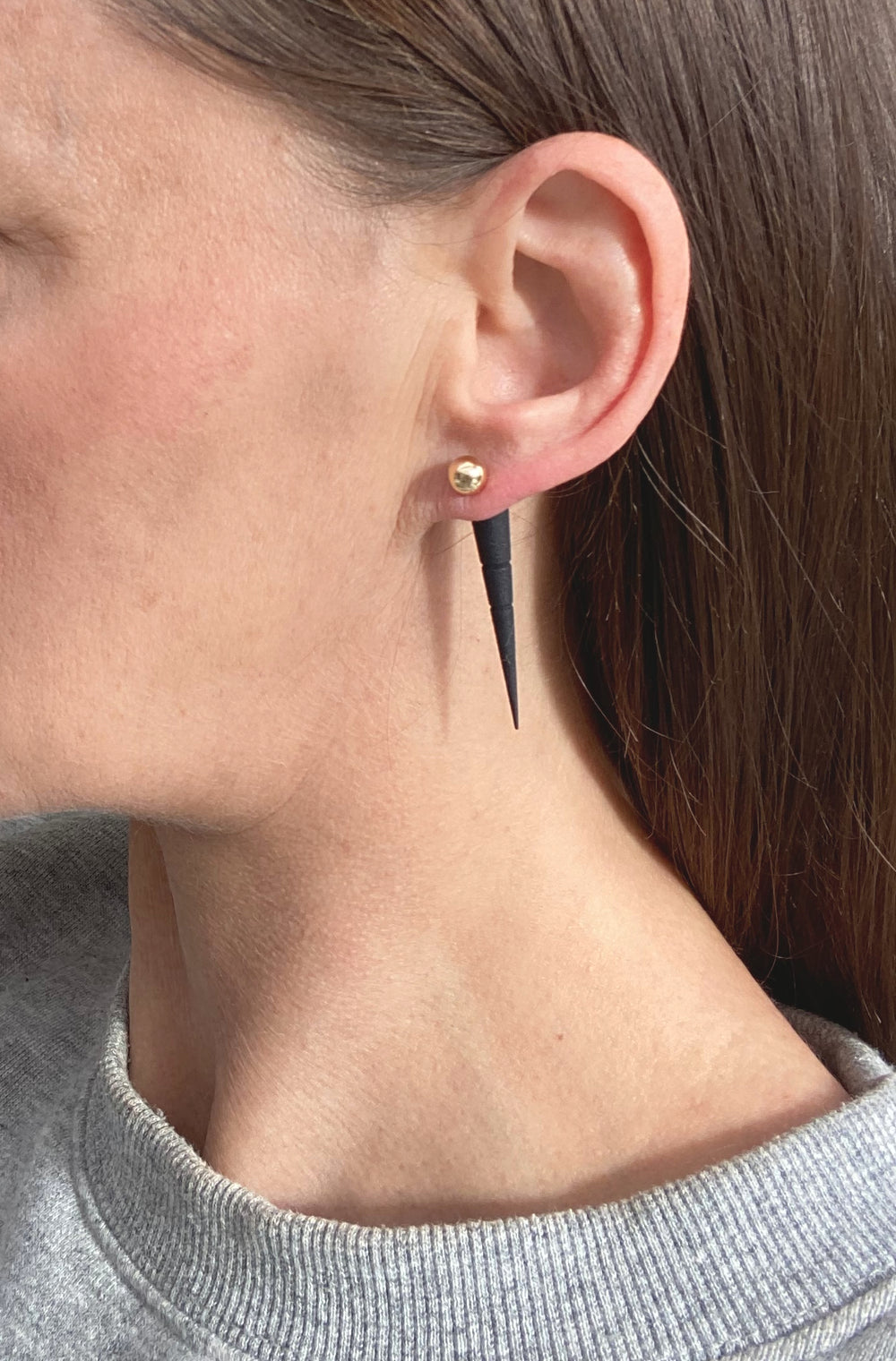 Close-up of a person with straight brown hair in a grey crew-neck top wearing Elsiem’s Rapture Sabre Earrings: 14K gold filled studs featuring long, narrow black 3D printed spikes.
