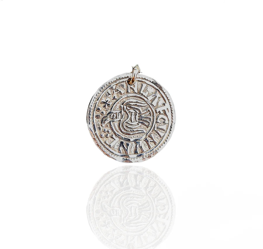 The Pale Raven "Raven Penny Viking Coin Charm" is a round sterling silver piece inspired by Viking coins, with engraved text and an abstract center, shown on a white background with a subtle reflection.