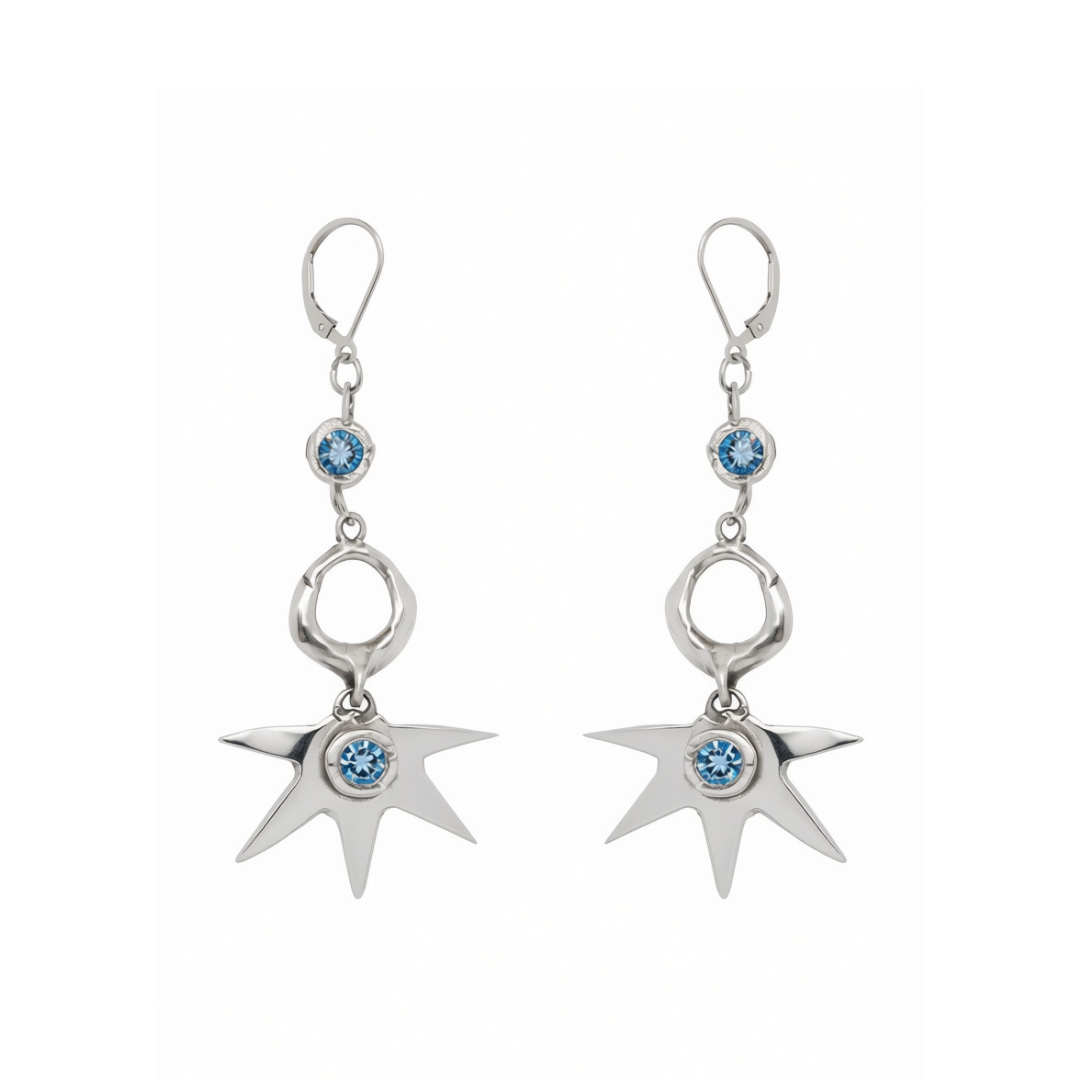 Réalta Earrings with 2 moissanite by Madeleine Blaine showcase circular and irregular silver forms adorned with blue gemstones, culminating in a striking starburst motif. Handcrafted to reflect the elegance of luxury jewelry, these earrings are beautifully presented against a white background.