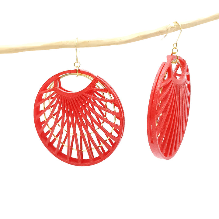 A pair of dangling Red Waterlily Earrings by Tochu-De with intricate red geometric designs hang from a light-colored branch. Crafted from 3D printed nylon jewellery, the earrings feature circular shapes with a pattern resembling radiating spokes. They are attached to gold hooks, and the background is plain white.