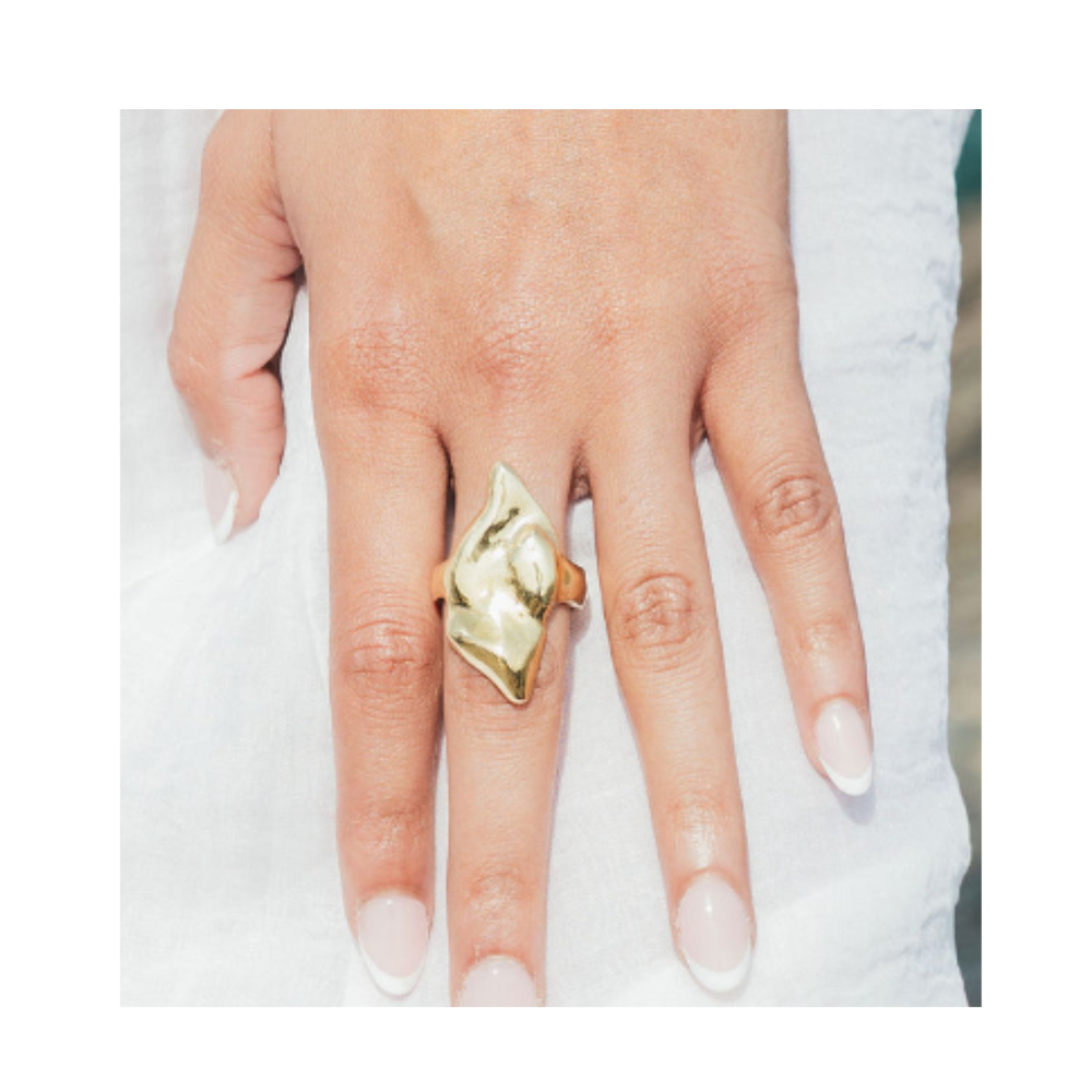 A hand with manicured nails wears the Rift Ring by Madeleine Blaine—an abstract, gold ring featuring striking negative space—resting on a white fabric background. Ideal for those who appreciate unique jewelry.