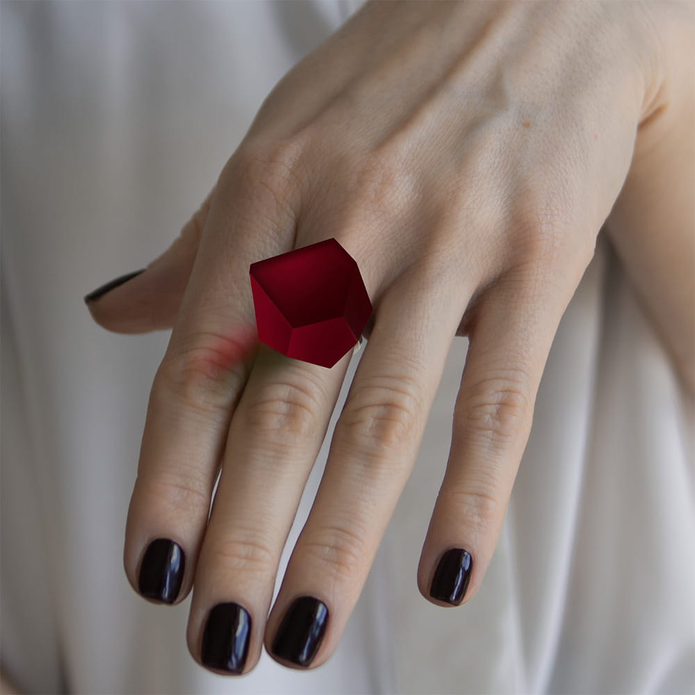 A hand with dark purple nail polish wears the Fruit Bijoux Ring Top VU Crystals Cherry Red by Fruit Bijoux on the middle finger. The softly blurred background and light garment highlight the ring's bold geometric design and vivid VU crystals.