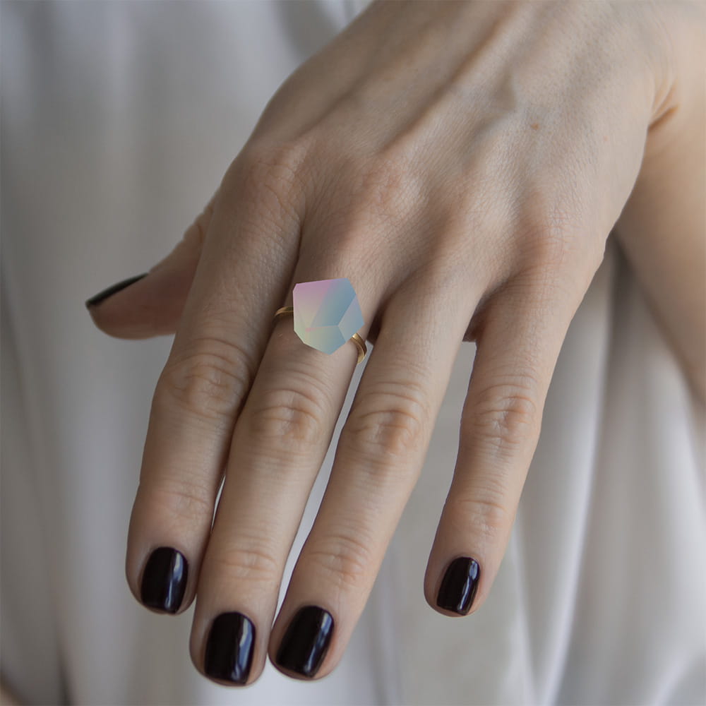A hand with dark purple nail polish wears the Fruit Bijoux Ring Top Unicorn by Fruit Bijoux, a gold-plated silver ring featuring a large, faceted pastel gemstone. A white garment can be seen in the background.