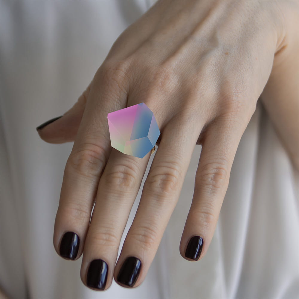 A hand with dark purple nails wears the Fruit Bijoux Ring Top Unicorn by Fruit Bijoux, featuring a large iridescent geometric gemstone set in sterling silver. The person is dressed in a white garment.
