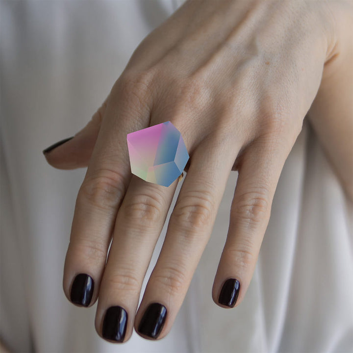 A hand with dark purple nails wears the Fruit Bijoux Ring Top Unicorn by Fruit Bijoux, featuring a large iridescent geometric gemstone set in sterling silver. The person is dressed in a white garment.