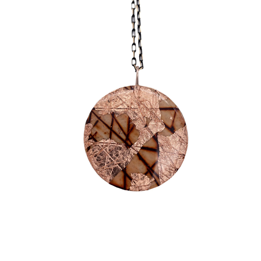 The Rothlú Ró Sgraffito Necklace in Rust/Rose Gold features a round, hand-patterned pendant with geometric design, copper foil accents, and dark lines on a faceted nickel-free chain against a white background.