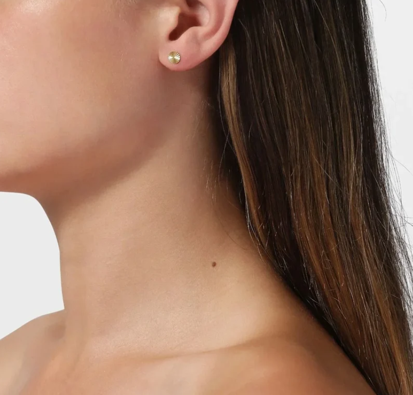 A close-up of a model's ear wearing a small gold stud earring set against a clean white background.