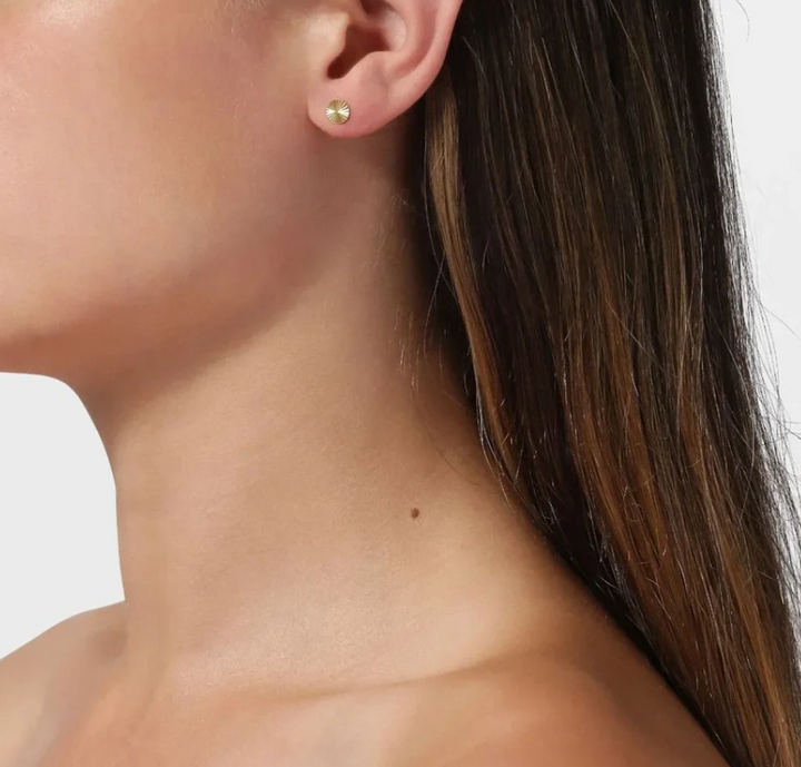 A close-up of a model's ear wearing a small gold stud earring set against a clean white background.