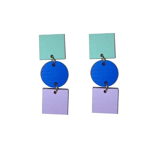The Ruth Earrings by Shock of Grey feature hand-painted birchwood shapes—green square, cobalt blue circle, and lilac square—linked by metal rings. Arrives in a recycled gift box.