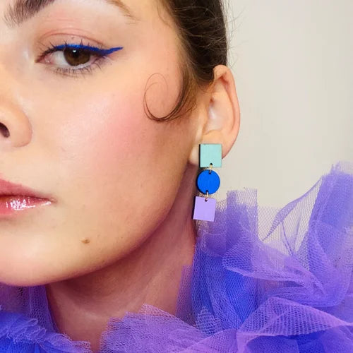A close-up of a person wearing bold blue eyeliner, glossy lips, and Shock of Grey's Ruth Earrings in Cobalt Blue, Lilac, and Duck Egg. They are dressed in a purple tulle garment with soft lighting and a neutral background.