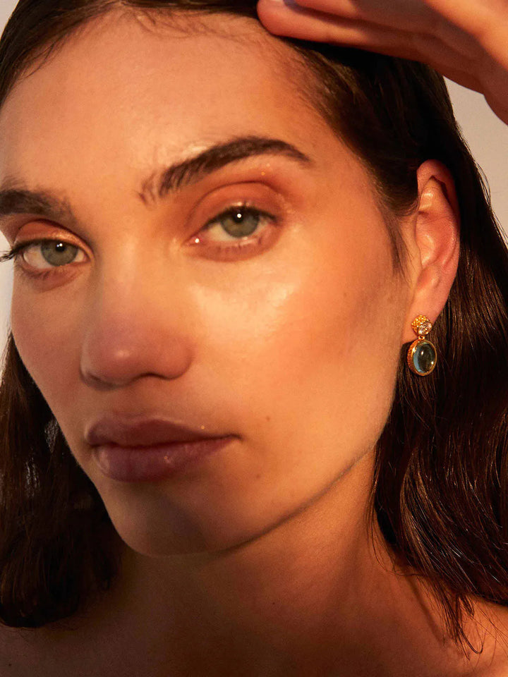 A close-up of a person with fair skin and brown hair wearing Serena Earrings by Shyla, crafted from 22-carat gold with a hand-hammered setting and featuring a dark gemstone. The image highlights their glowing skin with natural makeup, focusing on their expressive eyes and the stylish earring. The background is neutral and softly lit.