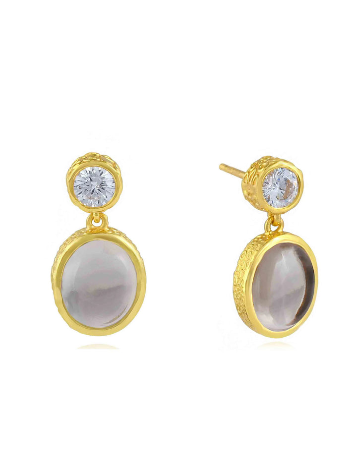 Introducing the Serena Earrings by Shyla: a pair of elegant earrings featuring a two-tier design. Each earring has a round, clear gemstone at the top connected to a larger, oval-shaped, greyish stone encased in a hand-hammered 22-carat gold setting. The smaller stone is a stud, while the larger one dangles below.